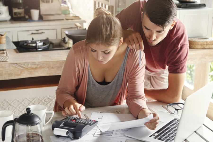 Family budget and finances concept. Young serious wife and husband doing accounts together at home, planning new purchase: woman holding document and making necessary calculations on calculator